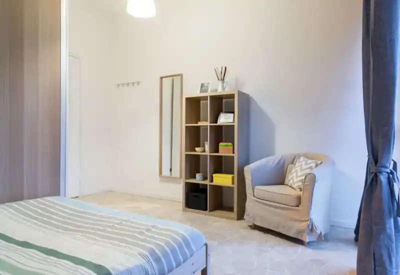 Bright, simply furnished bedroom with a visible double bed, a comfy armchair, and open shelf storage — neutral tones and soft lighting create a calm atmosphere.
