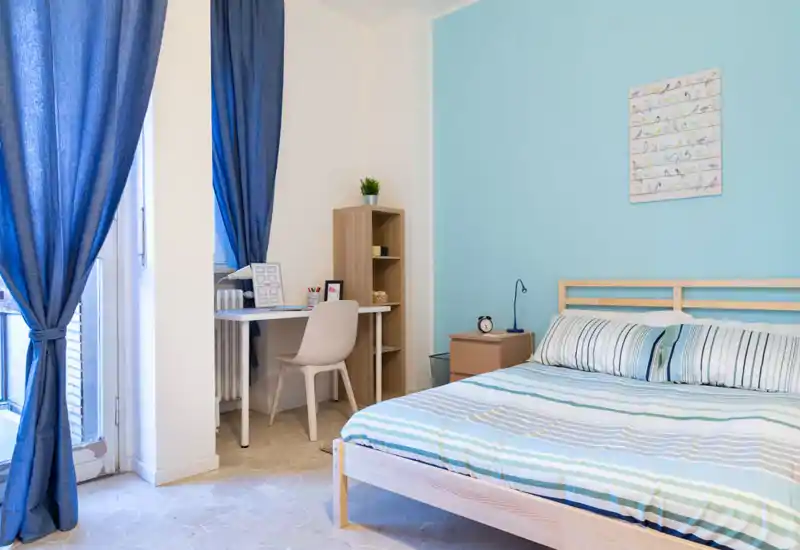 Bright, airy bedroom with a wooden bed, striped bedding and soft blue accent wall; a small desk by the window creates a practical work corner.