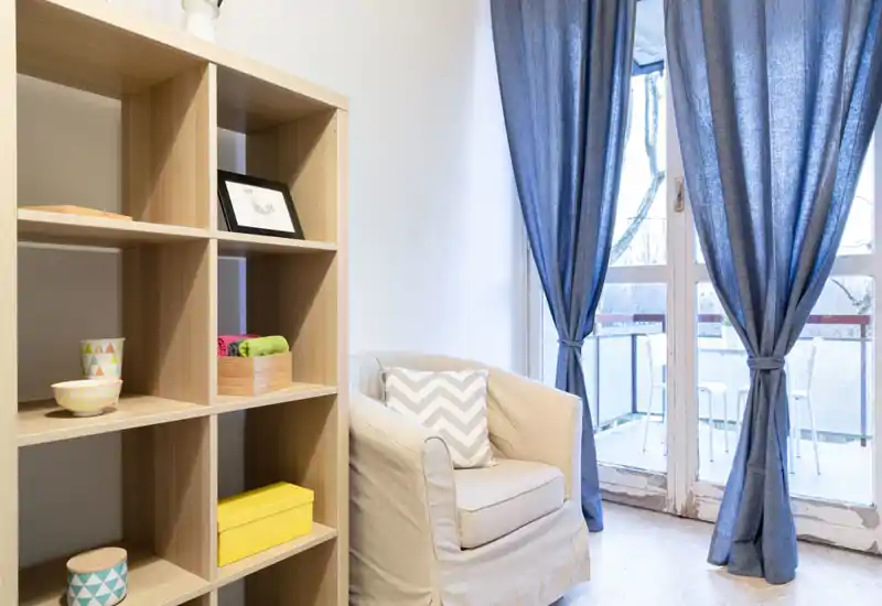 Bright, cozy living area with a comfortable armchair, decorative shelving and natural light from the balcony doors — ideal for relaxing.