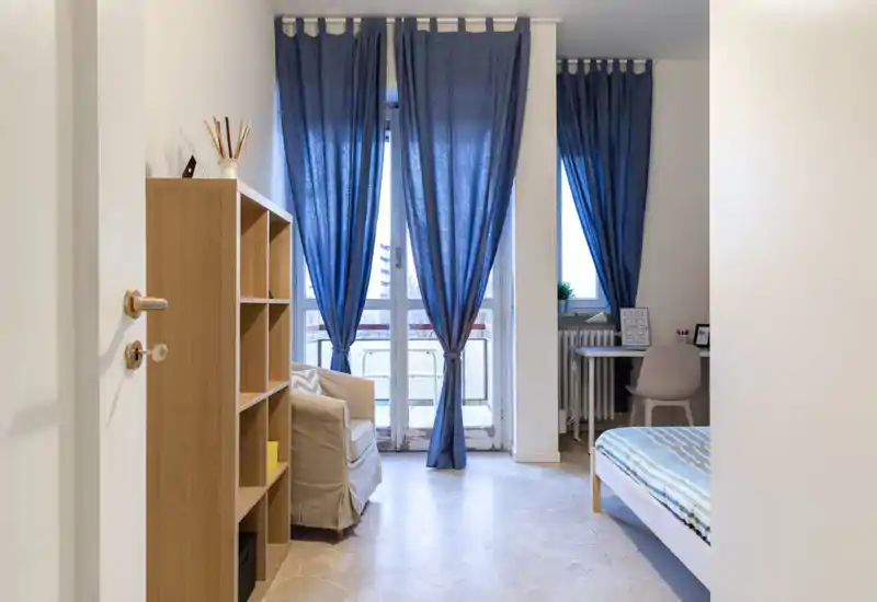 Bright, tidy bedroom with a visible bed, a cozy armchair and large windows dressed with blue curtains letting in natural light.