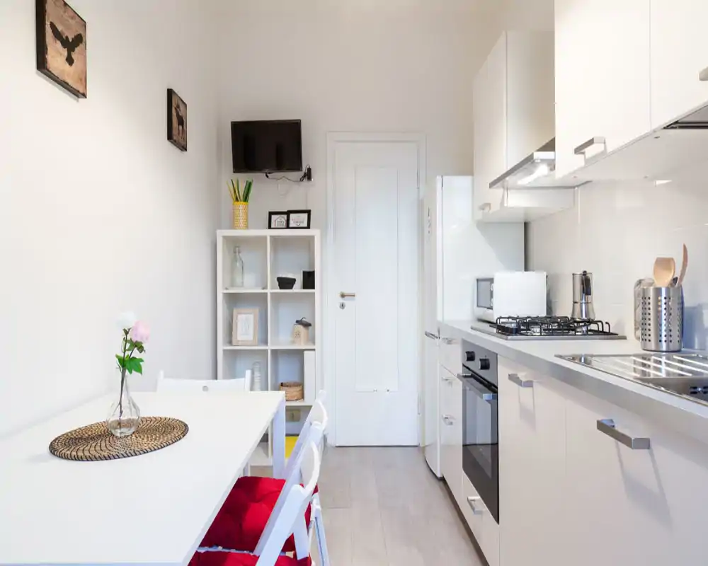 Bright, compact kitchen with integrated dining area: white cabinets, gas hob, oven and a small table with seating and decorative touches.