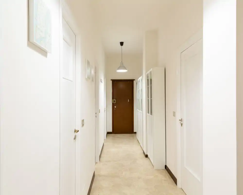 Bright, minimalist entrance corridor with white walls, stone floor and a wooden front door; fitted cabinets provide storage and a centered pendant light illuminates the passage.
