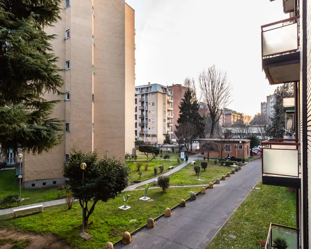 Communal outdoor courtyard with paved walkway, small lawn areas, trees and surrounding apartment buildings—pleasant view from a balcony suitable for listing the exterior space.