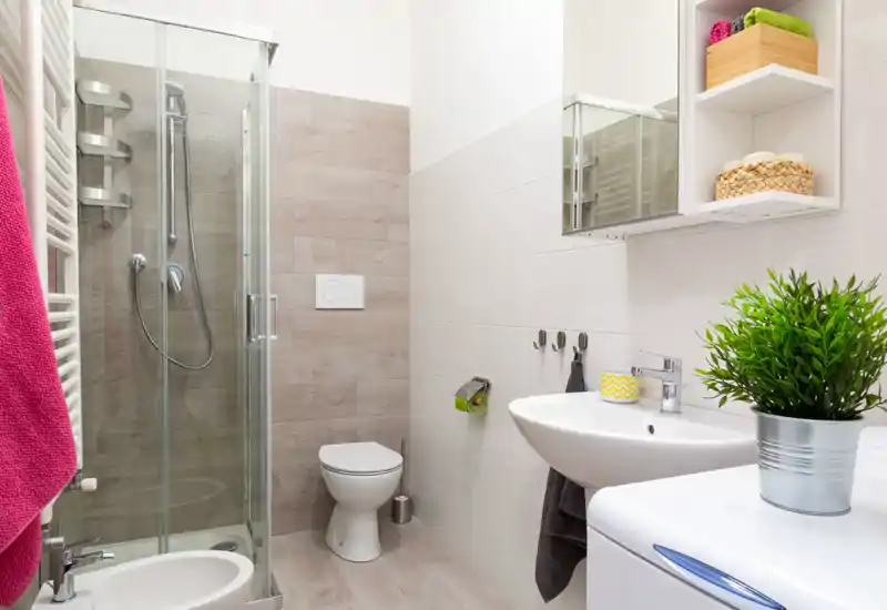 Bagno moderno e luminoso con box doccia in vetro, wc a parete, lavabo e mensole per riporre gli oggetti; asciugamani colorati e una piantina creano un aspetto accogliente.