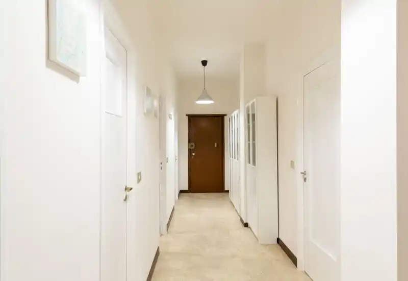 Ingresso luminoso e pulito con pareti bianche, pavimento in piastrelle e una lampada a sospensione centrale che conduce a una porta d'ingresso in legno. Armadiature sul lato offrono spazio di contenimento, perfetto come ingresso accogliente.