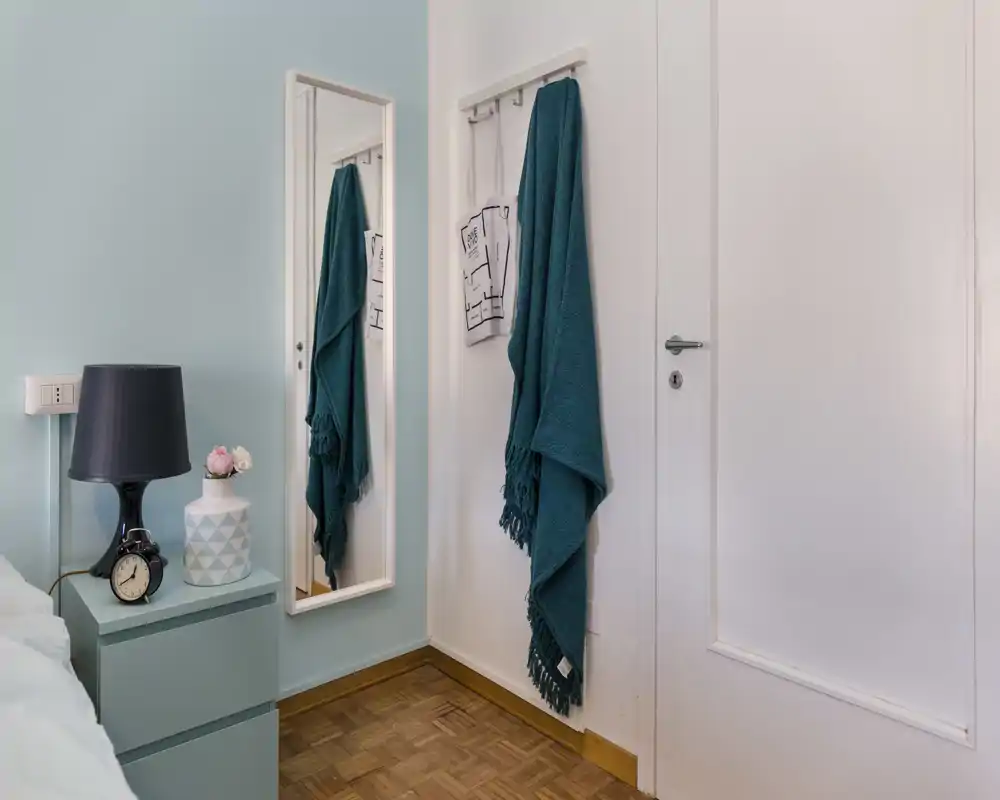 Small, stylish bedroom corner with a bedside table, lamp, decorative vase and a teal throw hanging on the wall; a mirror reflects the room and a bed edge is visible at left.