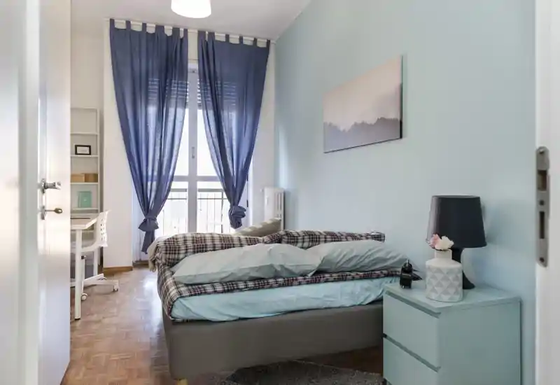 Bright, stylish bedroom with a comfortable double bed, bedside table and natural light from a balcony window. Soft blue walls and coordinated textiles give a calm, welcoming atmosphere ideal for rental listings.