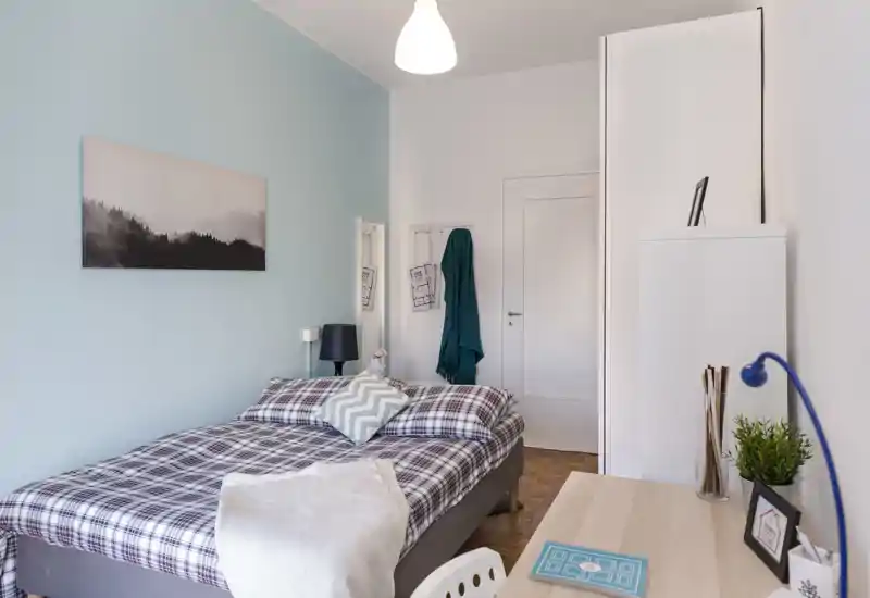 Bright, neatly staged bedroom with a double bed dressed in plaid linens, a bedside lamp, small desk and calming pastel wall color — ideal for rental photos highlighting comfort and style.