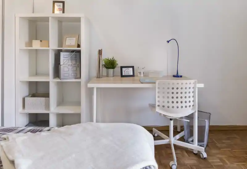 Bright, minimalist bedroom corner with a visible bed in the foreground, a white desk with a blue lamp and a shelving unit for storage — ideal for a cozy single or guest room.