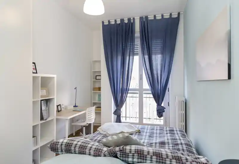 Bright, well-staged bedroom with a double bed dressed in plaid bedding, a workspace with shelving, and a large window with blue curtains letting in soft natural light.