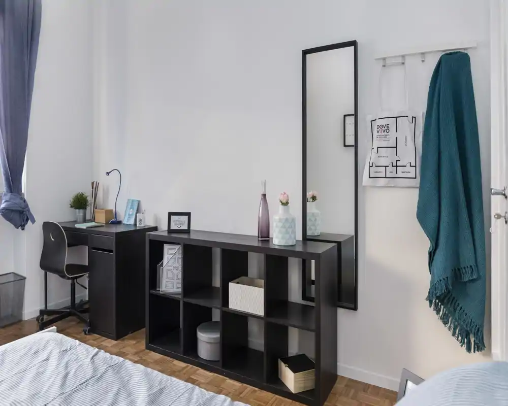 Bright, well-styled bedroom corner with a visible bed, a desk by the window and open shelving; modern decorative vases and a full-length mirror create a tidy, inviting space.