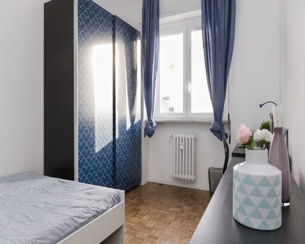 Bright, tidy single bedroom with a visible bed, patterned blue wardrobe and a desk by the window. Natural light and coordinated blue textiles create a fresh, welcoming atmosphere.