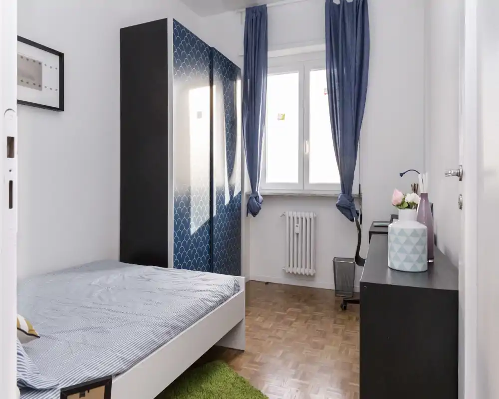 Bright, neatly styled bedroom with a single bed, patterned wardrobe and desk by the window — clean parquet flooring and blue curtains create a calm atmosphere.