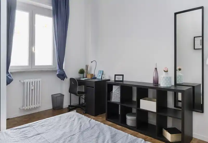 Bright, modern bedroom with a visible bed in the foreground, a study desk by the window and a low shelving unit with decorative objects — clean, minimal styling and abundant natural light.