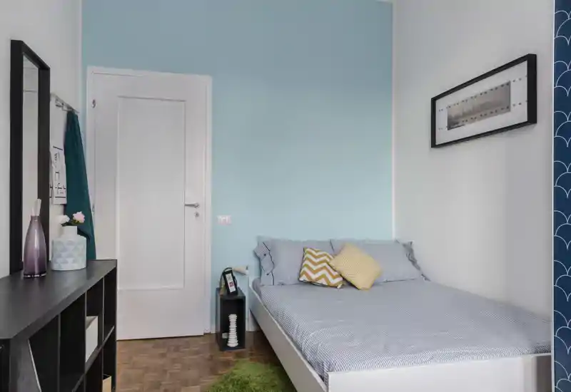 Well-lit, modern single bedroom with a neatly made bed, decorative cushions and a compact bedside shelf. Soft blue accent wall and coordinated textiles create a calm, inviting atmosphere.