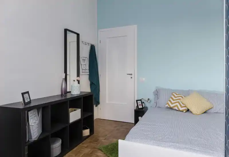 Bright, tidy bedroom with a striped bed, chevron and dotted accent pillows, a bedside table and a low shelving unit — calm blue wall and neat, modern styling.