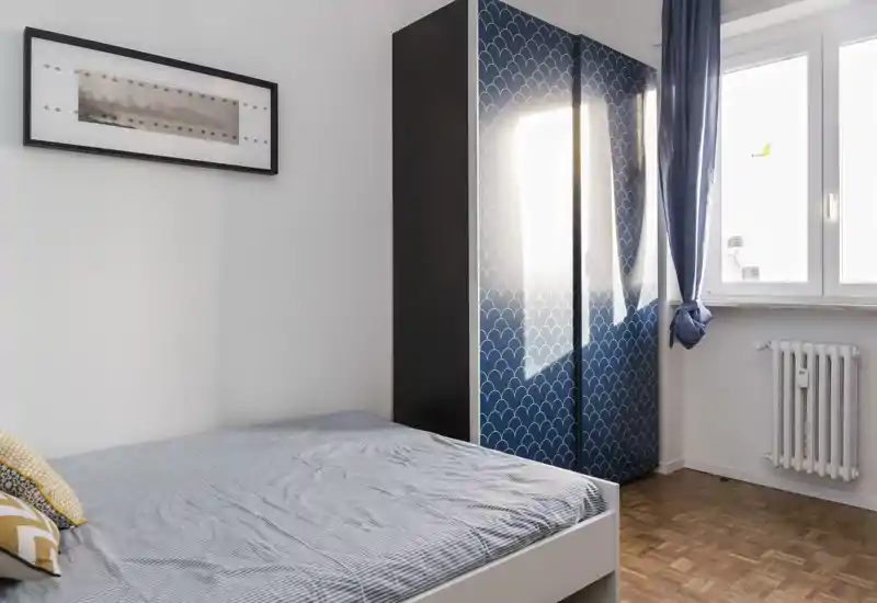 Bright, modern bedroom with a neatly made double bed, patterned blue wardrobe and natural light from a window.