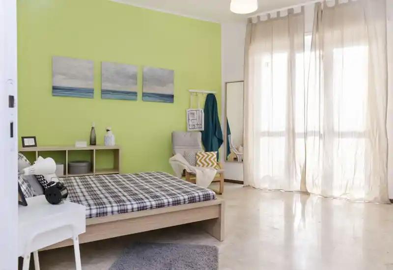 Bright, modern bedroom with a wooden low bed, patterned bedding, a cozy armchair and large sheer-curtained windows that fill the room with natural light.