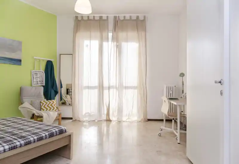 Bright, simply furnished bedroom with a single bed, armchair and a small desk by the window. Sheer curtains diffuse natural light and the green accent wall adds a fresh touch.