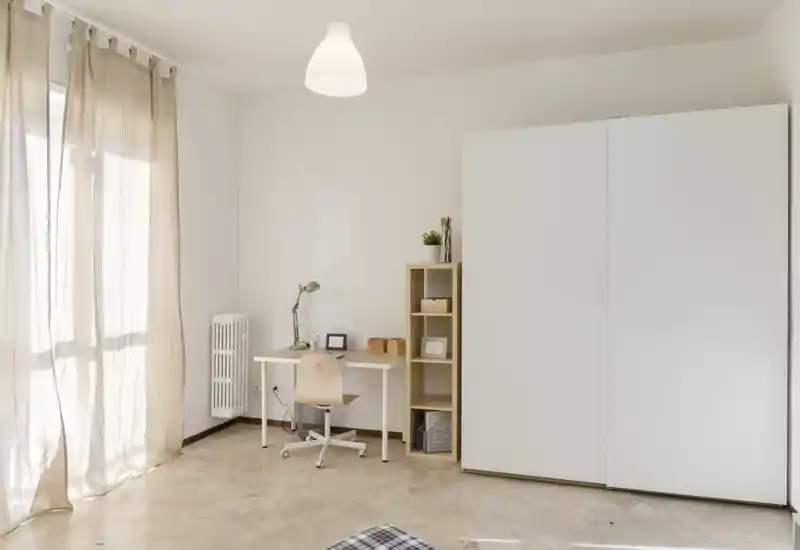 Bright, minimalist bedroom corner with a large white wardrobe, small desk and natural light from a sheer-curtained window.