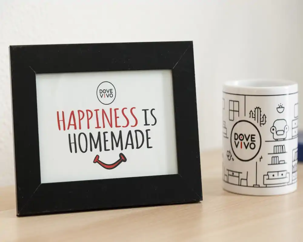 Gros plan d'un cadre indiquant « HAPPINESS IS HOMEMADE » et d'une tasse de marque sur une surface en bois ; image décorative adaptée au lifestyle/branding, elle ne montre pas une pièce identifiable.