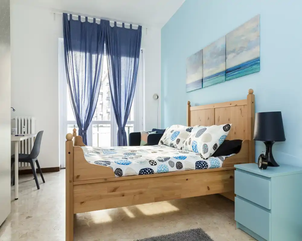 Chambre lumineuse et soignée avec un lit double en bois, une literie à motifs et des touches de bleu coordonnées. Grande fenêtre avec rideaux bleu marine laissant entrer la lumière naturelle, table de chevet avec lampe fonctionnelle.