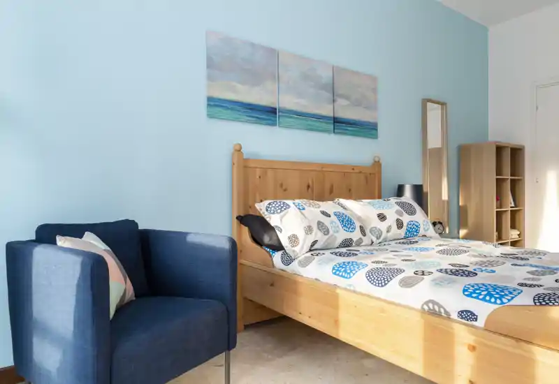 Camera da letto luminosa e ben arredata con letto matrimoniale in legno, biancheria a fantasia e una poltrona blu. Quadri a tema mare e pareti azzurro chiaro donano un’atmosfera fresca e rilassante.