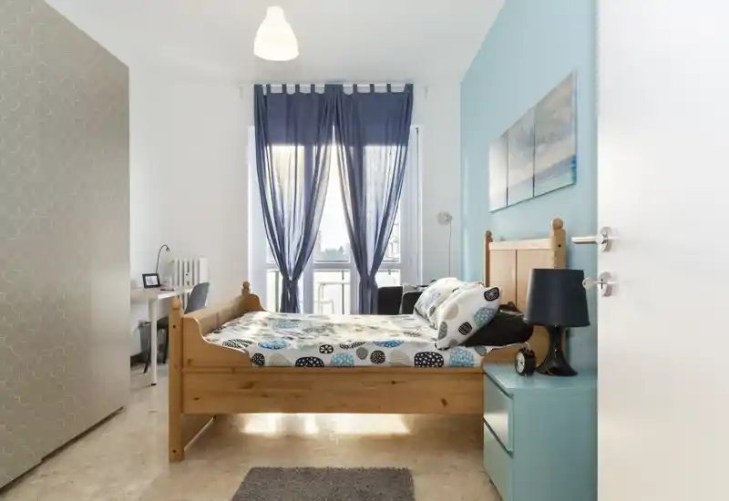 Camera da letto luminosa e ben arredata con letto in legno, biancheria a fantasia, comodino e scrivania vicino alla finestra. La luce naturale filtrata dalle tende blu crea un'atmosfera tranquilla e accogliente.