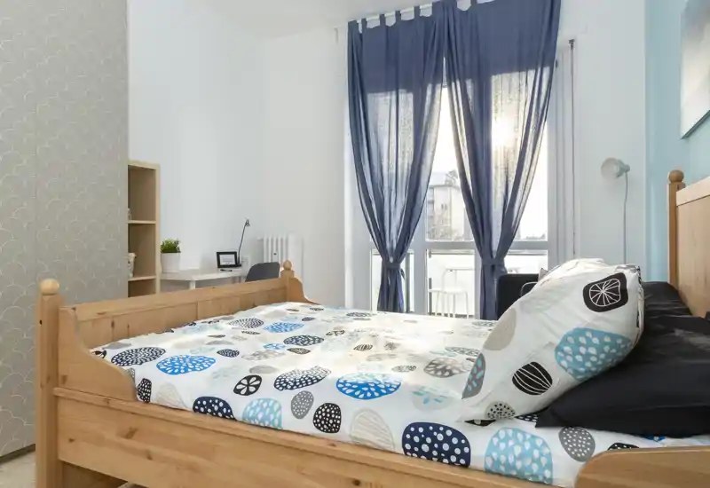 Camera da letto luminosa e ordinata con un letto matrimoniale in legno in primo piano, copriletto decorato e una scrivania accanto a una grande finestra che lascia entrare luce naturale.