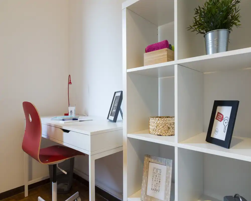 Angolo studio luminoso e ordinato con scrivania bianca e sedia rossa accanto a una libreria a cubi con oggetti decorativi e una piccola pianta.