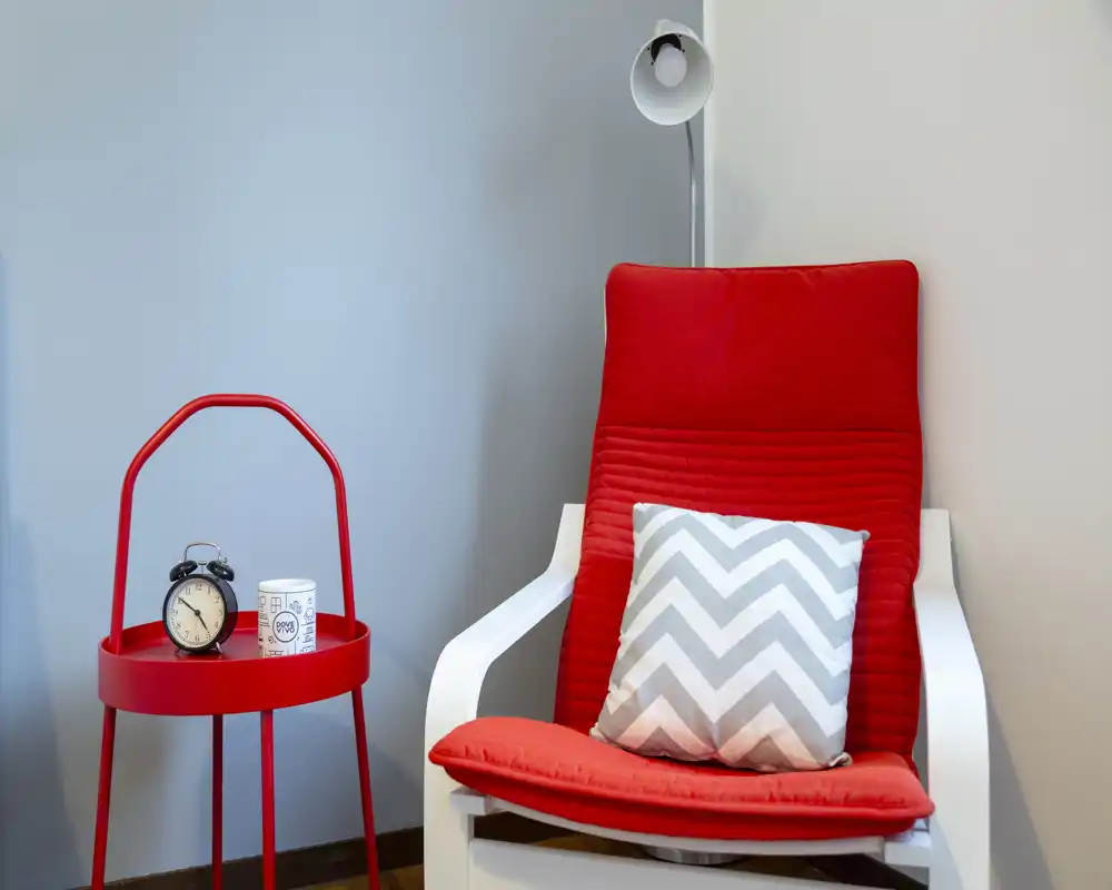Angolo lettura elegante e accogliente con poltrona rossa, cuscino a zig-zag e piccolo tavolino rosso con sveglia — moderno e luminoso.