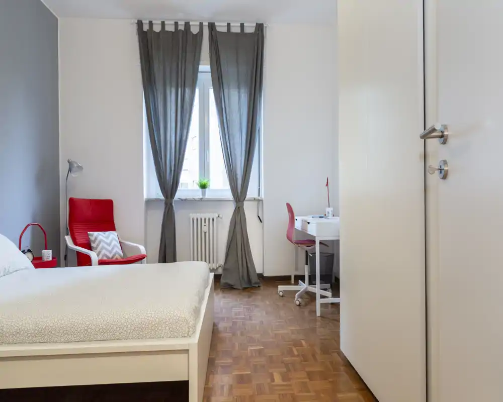 Camera da letto moderna e luminosa con letto bianco, pavimento in parquet, una poltrona rossa accogliente e una scrivania vicino alla finestra.