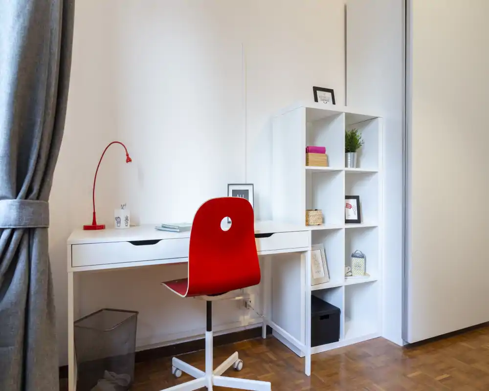 Angolo studio luminoso e ordinato con scrivania bianca, scaffale e sedia girevole rossa, completato da una lampada e decorazioni—ideale per concentrarsi.