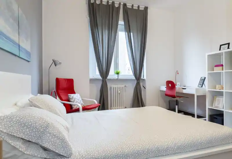 Camera da letto moderna e luminosa con comodo letto matrimoniale, poltrona rossa, scrivania e scaffalatura ordinata — ambiente ben curato ideale per rilassarsi o lavorare.