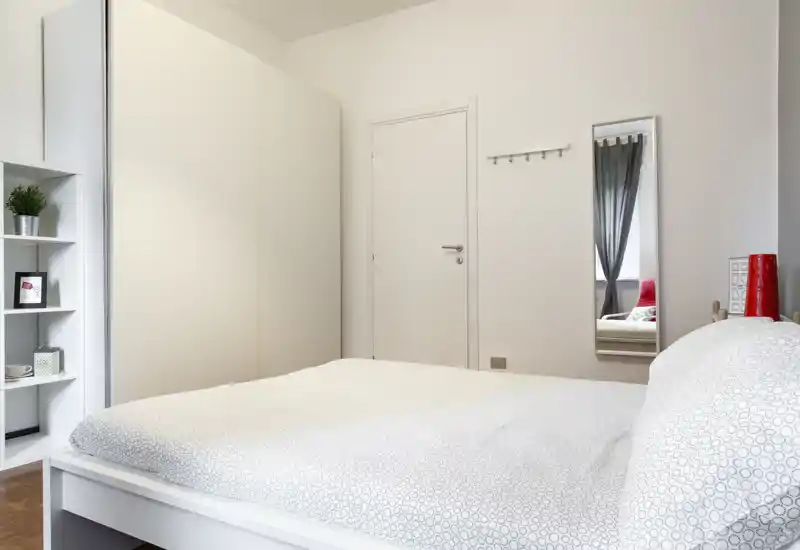 Camera da letto luminosa e minimal con un grande letto bianco in primo piano, un armadio alto e mensole a sinistra e uno specchio a figura intera sulla parete. Toni neutri e arredamento ordinato creano uno spazio tranquillo e accogliente.