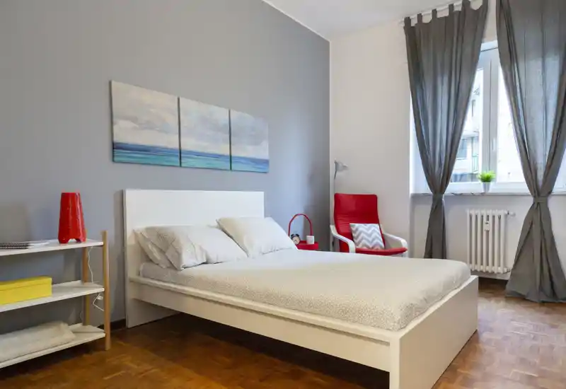 Camera da letto moderna e luminosa con letto bianco contenitore, biancheria a motivo, quadro a tre pannelli e una poltrona rossa vicino alla finestra per un angolo lettura accogliente.