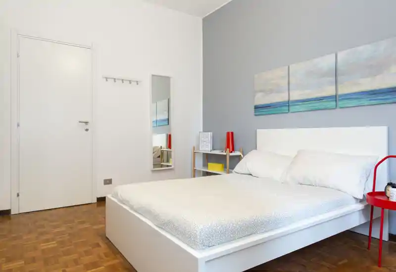 Camera da letto luminosa e moderna con un letto piattaforma bianco, biancheria semplice e arredo minimal. Un trittico con paesaggio marino sopra la testata e un piccolo comodino rosso per un tocco di colore.