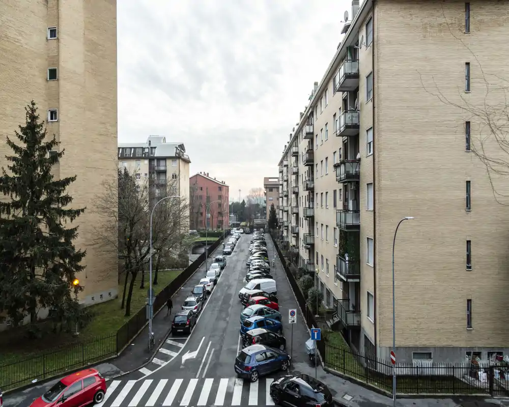 Veduta esterna a livello strada di un isolato residenziale con auto parcheggiate, attraversamento pedonale e palazzi di media altezza sotto un cielo coperto.