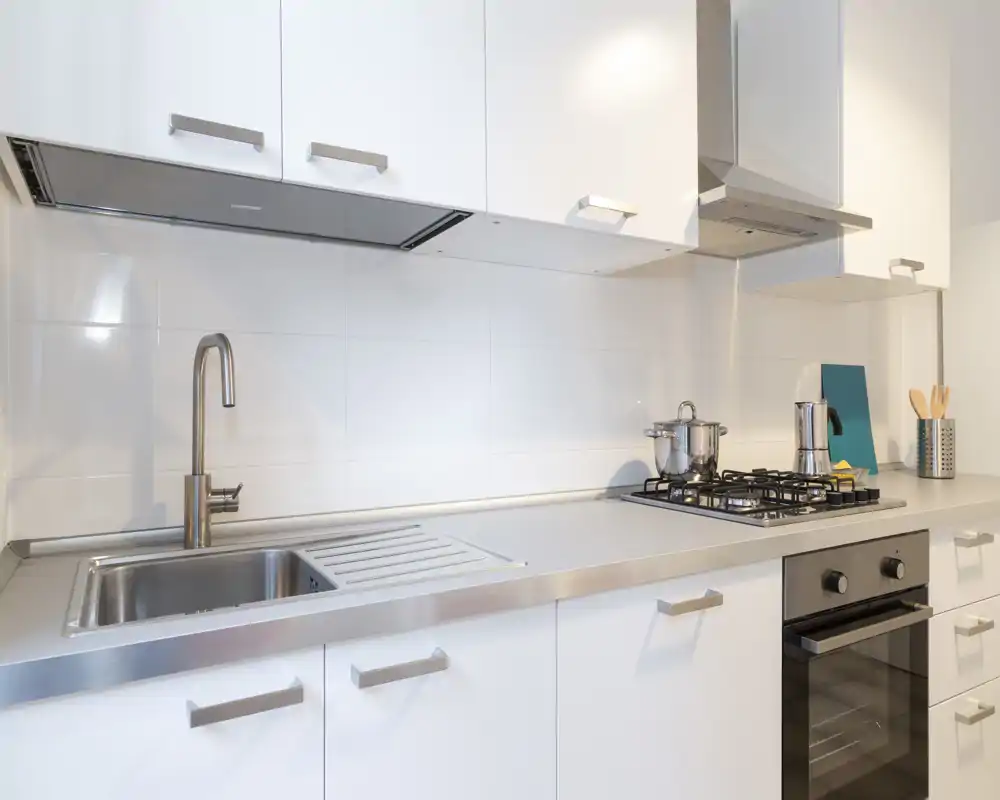 Cucina moderna e luminosa con mobili bianchi, piano in acciaio inox e piano cottura a gas con forno incassato — ambiente pulito e ben illuminato ideale per promuovere una kitchenette contemporanea.