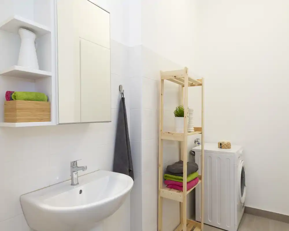 Bagno piccolo e luminoso con lavabo a muro, specchio con anta, appendino per asciugamano e scaffale in legno accanto a una lavatrice. Piastrelle bianche pulite e asciugamani colorati piegati donano un aspetto curato e accogliente.