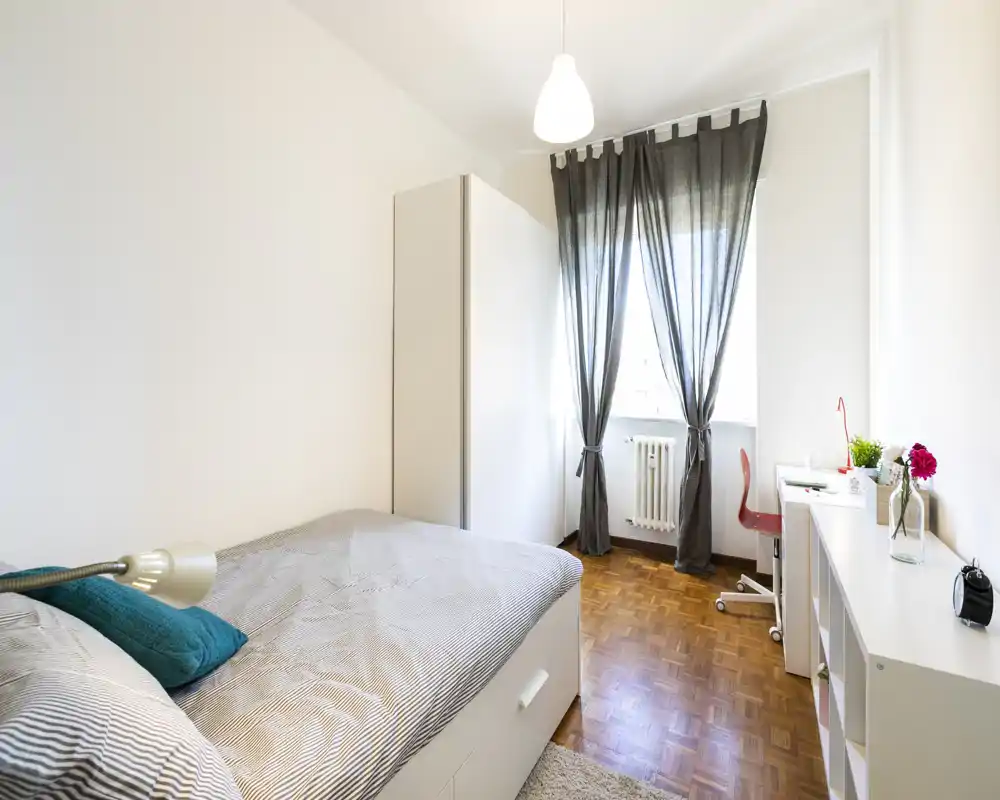 Camera da letto singola luminosa e ben arredata con letto comodo, armadio bianco e una scrivania vicino alla finestra. La luce naturale filtra dalle tende leggere creando un’atmosfera tranquilla e fresca, perfetta per annunci.