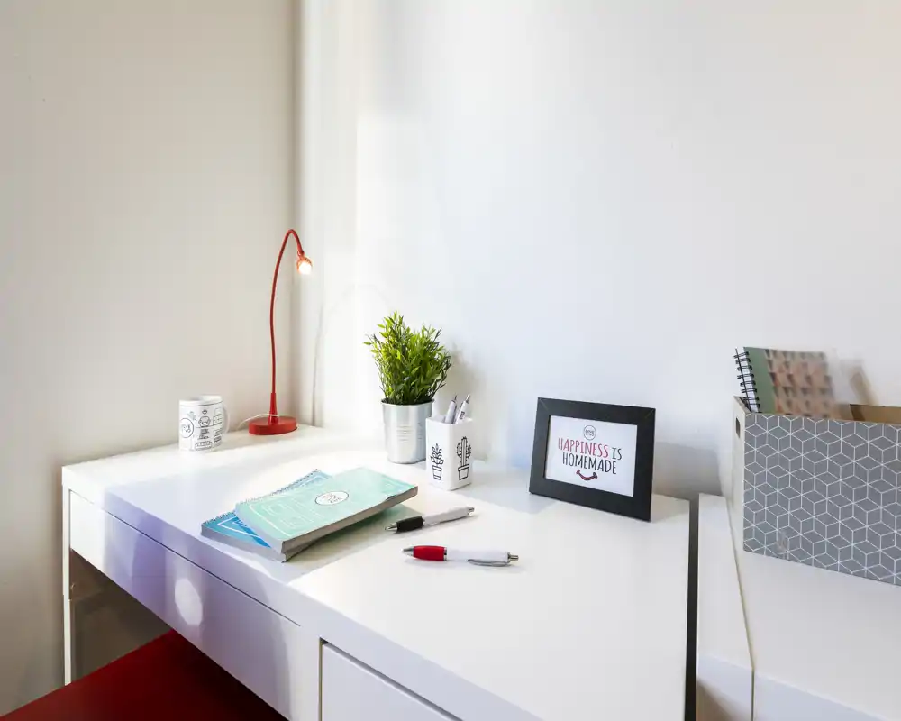 Angolo scrivania luminoso e minimal con una lampada rossa, una pianta in vaso e materiale di cancelleria su una superficie bianca — uno spazio di lavoro domestico ordinato ideale per concentrarsi.