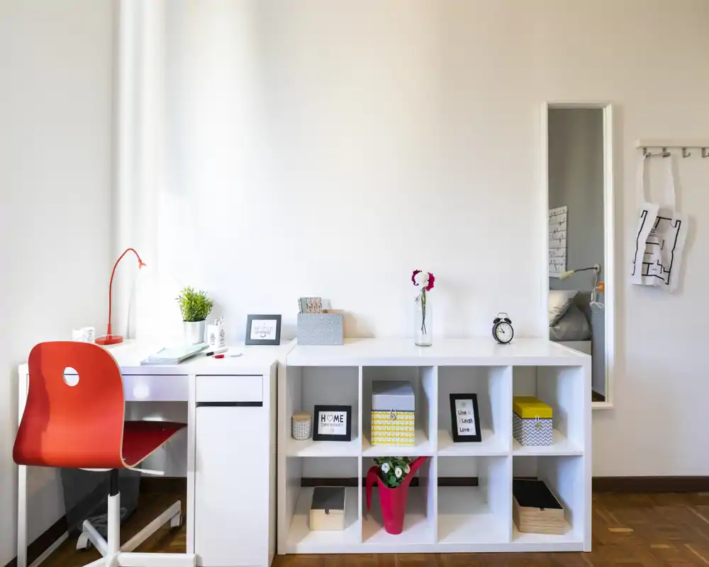 Angolo living/lavoro moderno e luminoso con mobile contenitore bianco, scrivania e sedia rossa; arredato con scatole e piante per un aspetto pulito e accogliente.