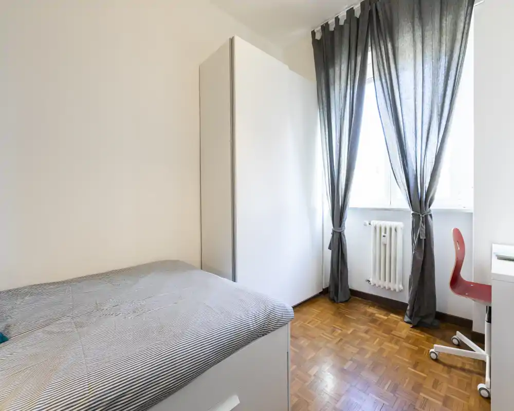 Camera da letto singola luminosa e minimal con letto rifatto, armadio bianco, pavimento in parquet e finestra con tende scure che lasciano entrare luce naturale.