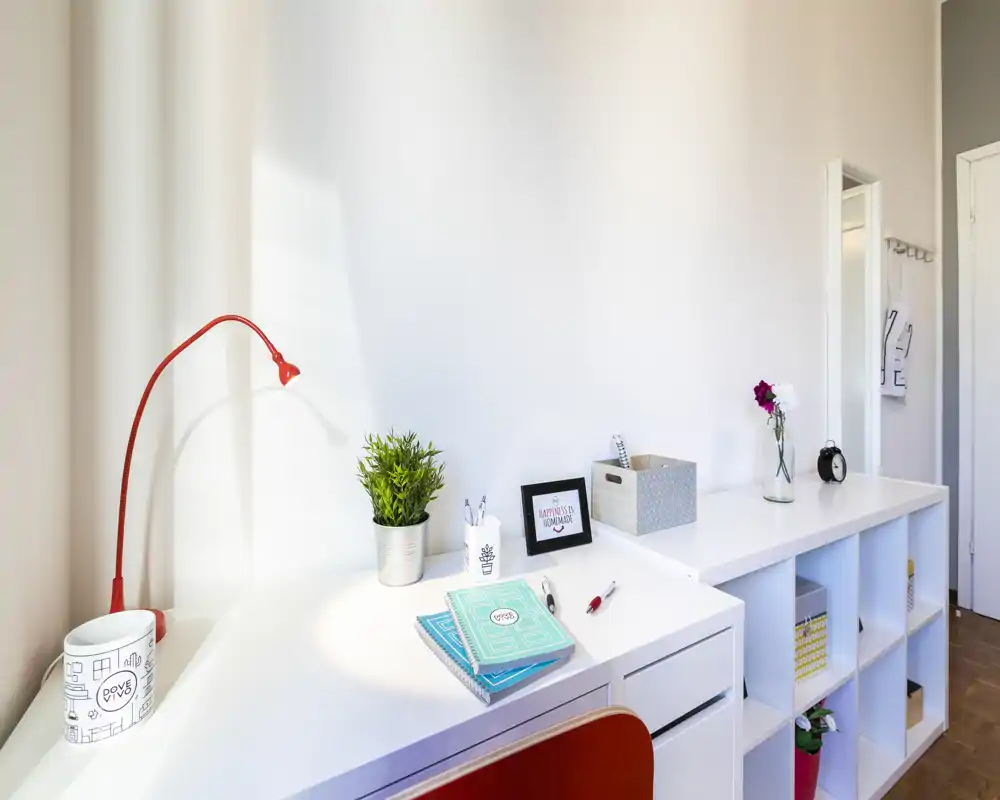 Area scrivania luminosa e moderna con piano bianco pulito, pianta decorativa, lampada da lavoro rossa e scaffalatura — perfetta per lavorare da casa.