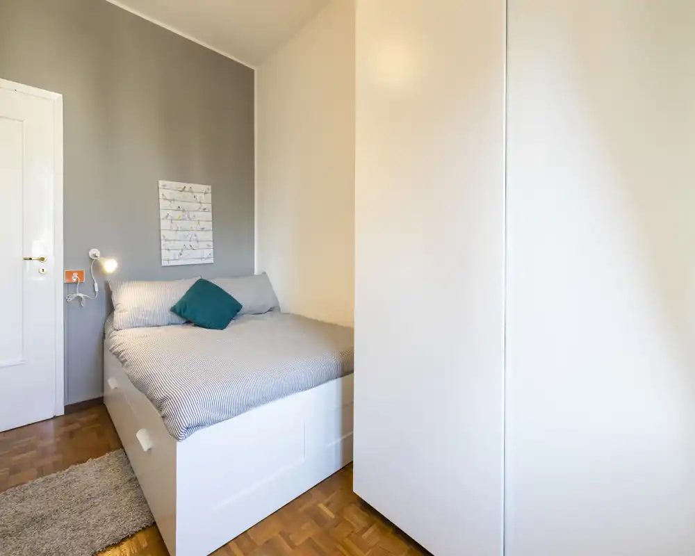 Camera da letto luminosa e ordinata con un ampio letto bianco a piattaforma, biancheria pulita e un cuscino verde acqua; pavimento in parquet e armadio integrato creano uno spazio compatto e accogliente.