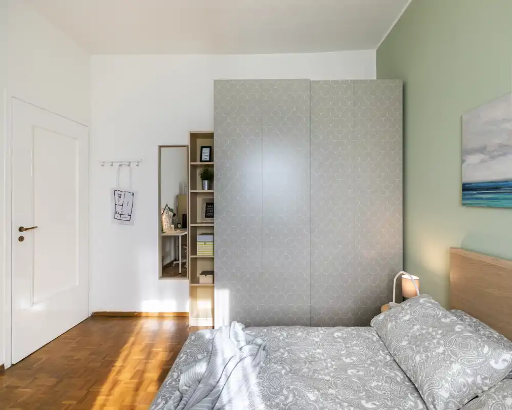 Comfortable, well-lit bedroom with a patterned wardrobe, bedside lamp and neat grey bedding; large bed and soft natural light make it ideal for listings.