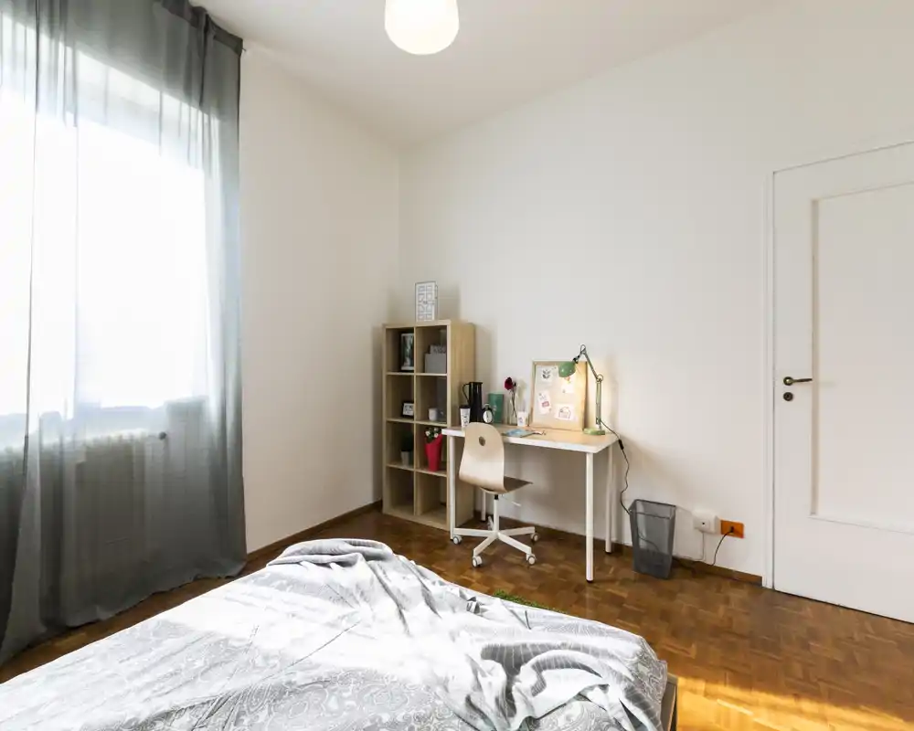 Bright, minimal bedroom with a visible bed in the foreground, a work desk and shelving by the wall, and soft natural light from a large window.