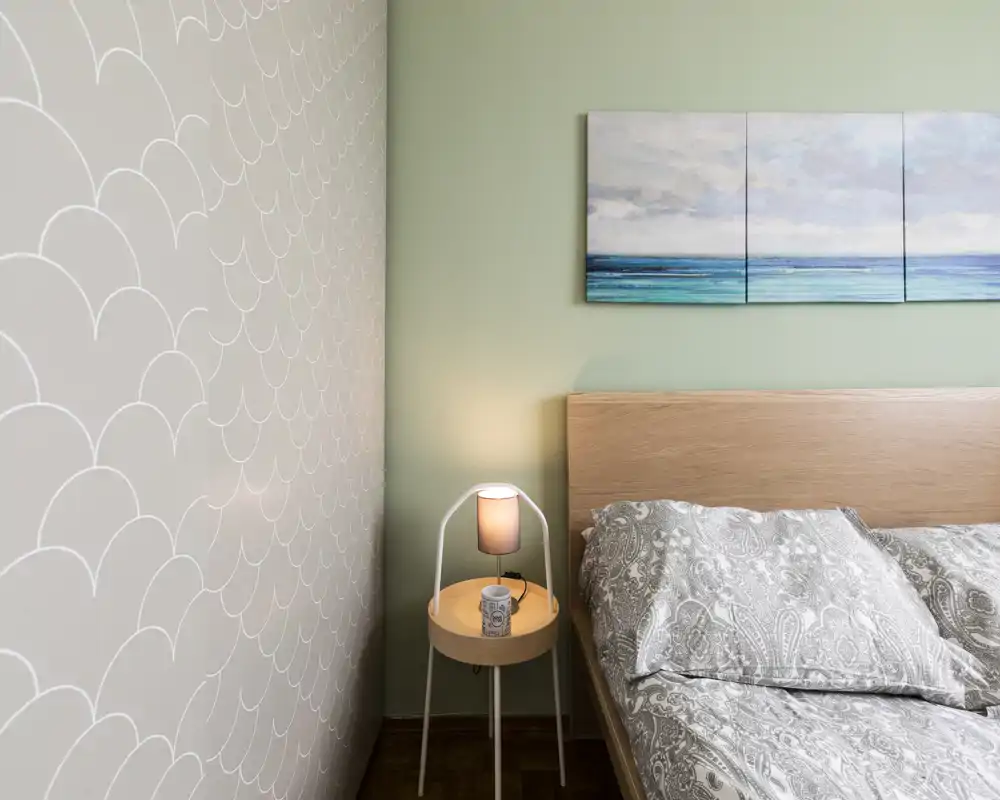 Comfortable bedroom corner with a wooden headboard, patterned bedding and a bedside table with lamp; soft green wall and coastal triptych add a calm atmosphere.