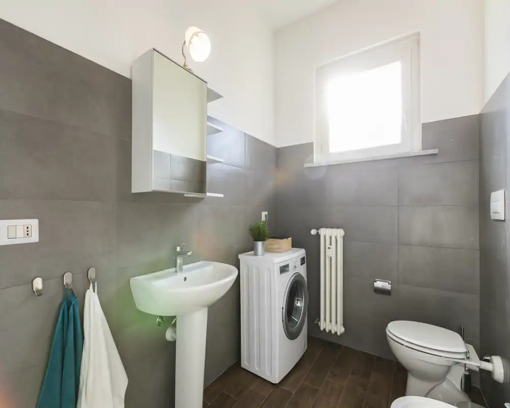 Bright, modern bathroom with pedestal sink, toilet and washing machine; grey wall tiles and wood-effect floor create a clean, functional look.